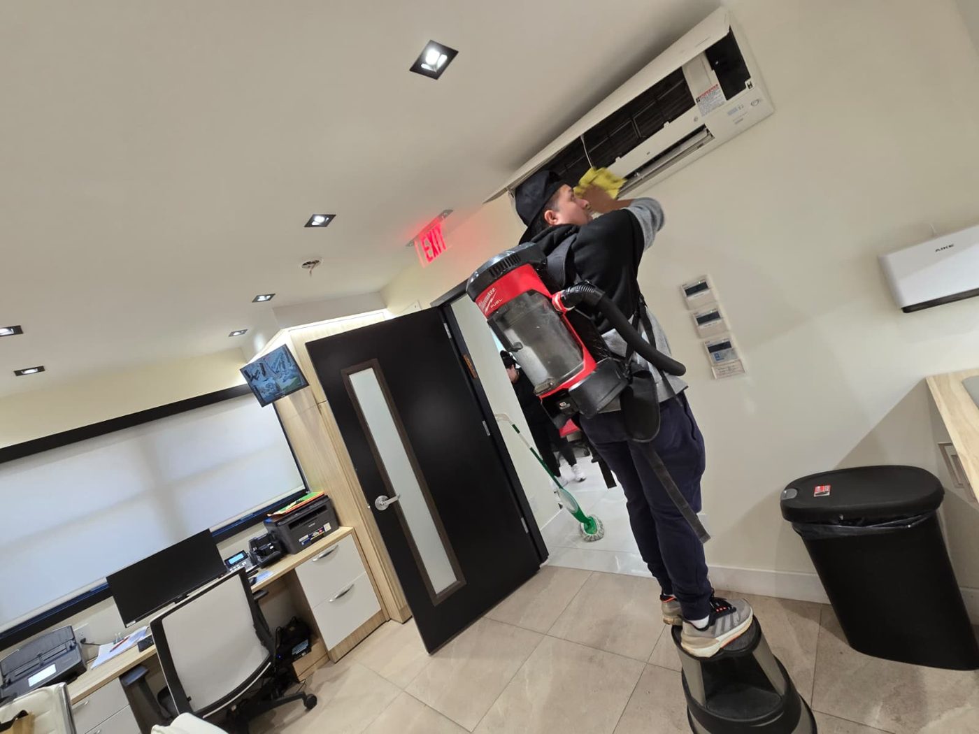 Fine Cleaning team member with a backpack vacuum wiping down a wall-mounted AC unit
